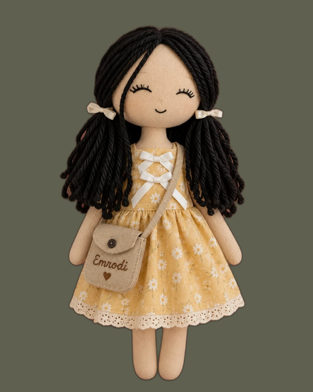 Doll with black yarn pigtails and cream bows in a yellow daisy dress with a lace hem, carrying a linen crossbody bag stitched with the name Emrodi